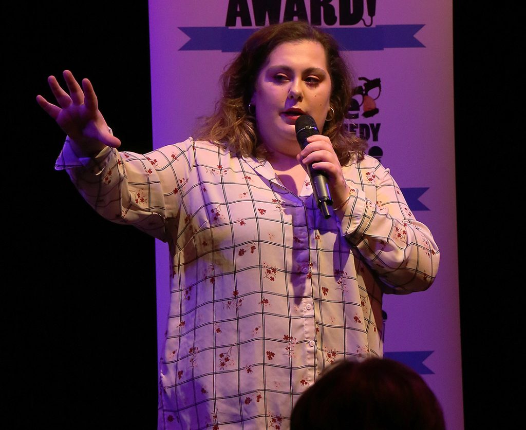 Maisy Whipp wearing a white checkered shirt with flowers on performing in front of the crowd. She is mid-speech and holding the microphone.