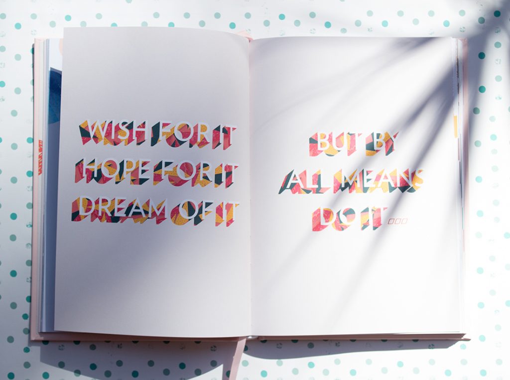 An artistic typography book laid out on a polka dot table that reads 'Wish for it, hope for it, dream of it. But by all means do it.' in orange and pink text.