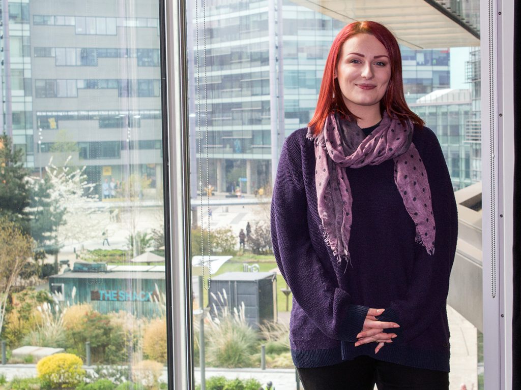 Hazel, who has red shoulder length hair and is wearing a dark purple jumper and a lighter purple scarf is stood in front of a wide window in our MediaCity building. Behind her you can see the communal garden that sits in the middle of MediaCityUK as well as the buildings behind her