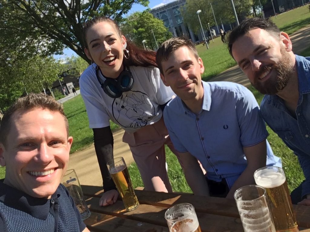 A photo of three men and a woman who has headphones round her neck. They are all sat at a picnic table on campus during a sunny day with beers in front of them. They are all smiling.
