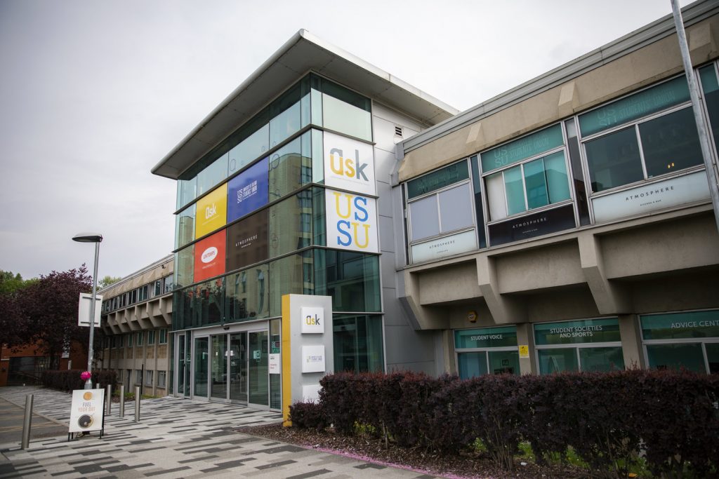 A campus shot of the University House also known as the University of Salfords' Students' Union. There is a row of bushes in front of it, as well as a signboard advertising the starbucks inside. There is also a sign for our AskUS service that has a grey and yellow colour palette.