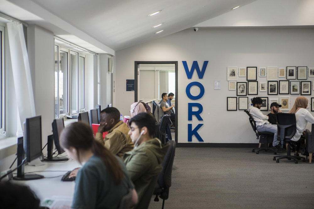 A shot of one of the computer areas in our Clifford Whitworth Library. The walls are freshly painted white but at the back you can see some art and photography framed on the wall next to students who are sat in black chairs at a table. The word 'Work' is fixtured on the wall in blue writing vertically. In the foreground you can see students sat at computers that are in front of windows.