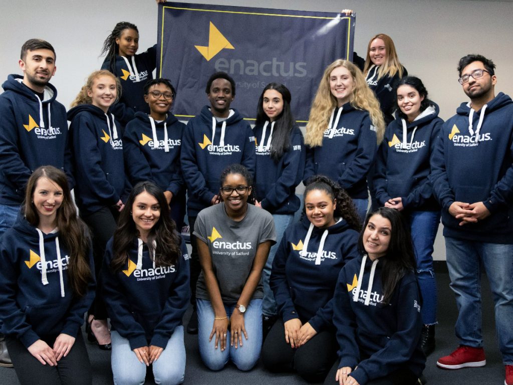 Members of the Enactus society all wearing navy blue hoodies with the 'Enactus University of Salford' logo on. Two students at the back of the group photo hold up a banner with the logo also on.