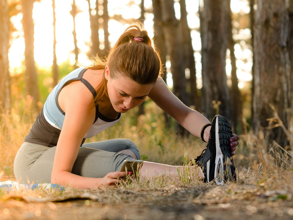 A woman with brunette hair in a ponytail wearing sportswear stretching on the ground in a forest. 
