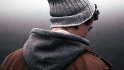 In the foreground of a gradient grey background stands a man with his back facing to the camera wearing a grey beanie and dark clothes.