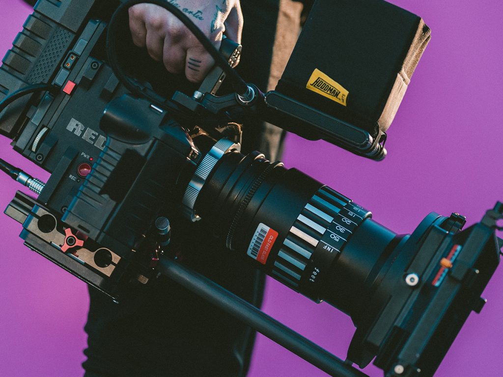 Against a bright purple background, a person with hand tattoos holds a large black camera video recorder