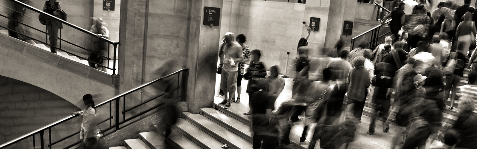 Edited in dark sepia/black and white this is a photo taken with adjusted shutter speed. It shows people and crowds moving throughout a station, people's movement is blurred.