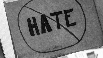 Taken or edited in black and white, someone is holding up a cardboard protest sign that shows the word 'HATE' crossed out to represent being against hate crimes