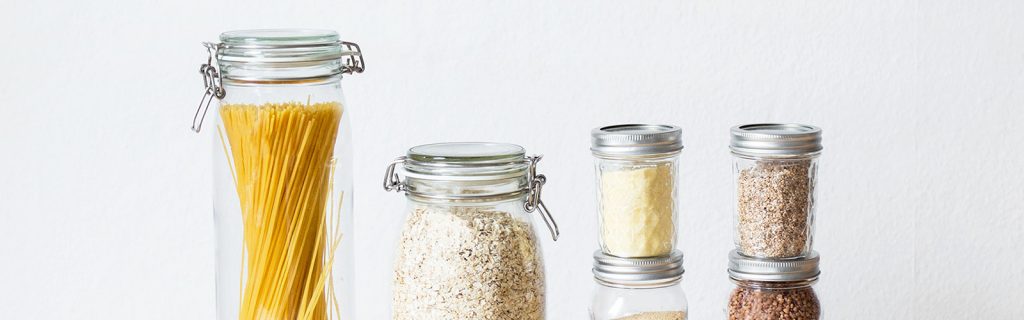 Image source is Photo by Laura Mitulla on Unsplash. It shows several glass jars with metal lids that have produce in it. On the left is a tall jar that has dry spaghetti in. The jar next to it seems to have oats inside it. This is against a white background.
