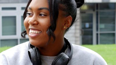 Artiana is sat in front of the New Adelphi building wearing a grey jumper and black headphones around her neck. She is looking into the distance and smiling.