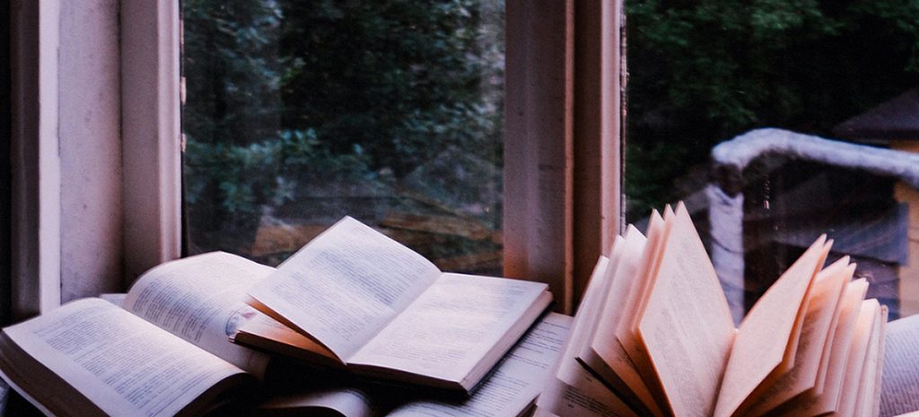 On a pale white windowsill there is a pile of opened hardback books scattered across the surface. The pages are different shades of clean white or slightly tinged with an beige colour from the sunlight. 