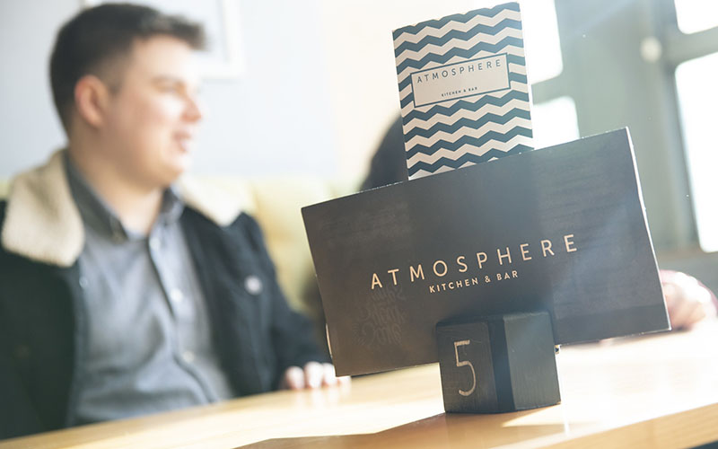 A photo taken at an angle that puts one of the table number stands at Atmosphere Kitchen and Bar as the main part of the shot. The table number is 5 and it's holding two menus that both read 'Atmosphere' on them. At the table in the background you can see a group of people sat chatting at the table. 