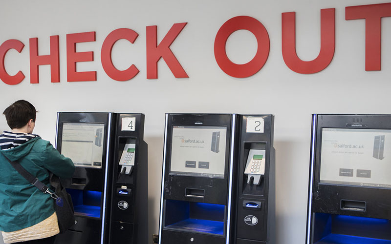 This photo is taken inside the Clifford Whitworth Library in the check out section on the ground floor. In the photo you can see three black self-check out screens that have square screens on above the scanner that you put your books in. These machines also have card readers on to pay for any loans or fees. Above the self-check outs on the white wall is a big red sign that just says 'Check out'. Someone in a green jacket is in the process of checking out books on the far left checkout stand.