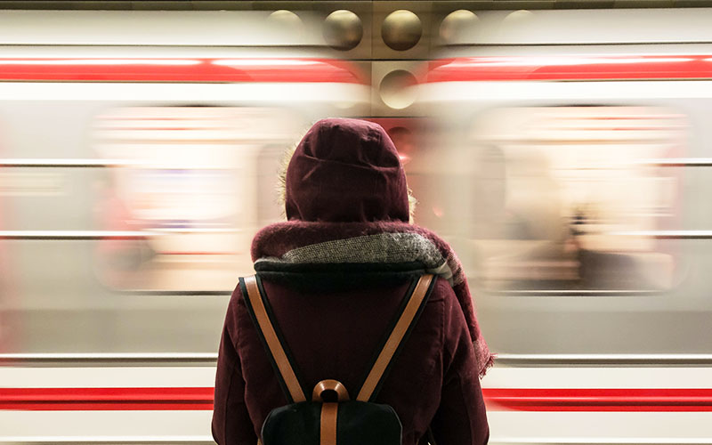 A young person with a petite frame wearing a dark red coat and knitted scarf is stood in front of a passing train. Using slow shutter photography the passing train is blurred. 