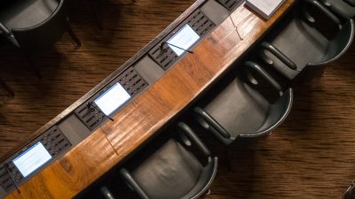An up-top shot of several black armchairs in front of wooden desks, set out like a meeting room or parliamentary discussion.