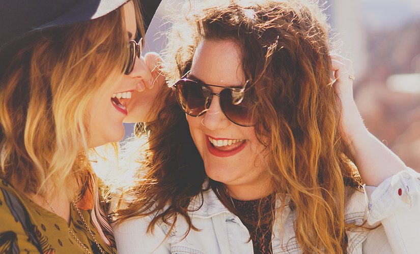 Two women one in sunglasses and one in sunglasses and a hat smiling and laughing together.