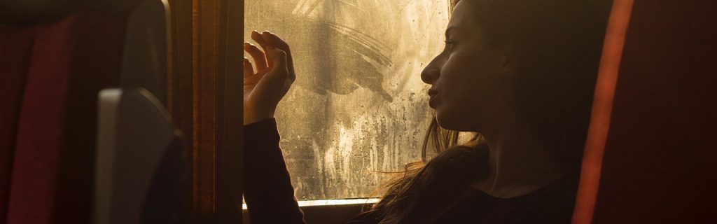 The photo, taken in lighting that gives the image a quite dark, evening atmosphere, shows a girl sat on some form of public transport that has red chairs. She is staring out of a window that has fogged up however you can see the mark where she has wiped it with her hand to look out of. 