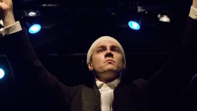 Performer Ryan O'Shea stands at the front centre of the stage, with a bandage wrapped around his scalp. He is holding a baton up as if to conduct an orchestra.