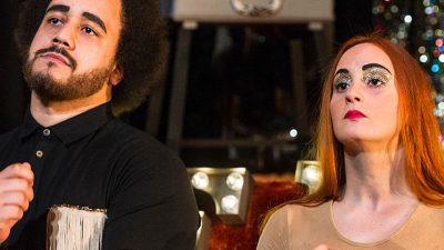 Bruno (left) and Sandy (right) sit on stage mimicking performing instruments. Bruno is playing the drums and Sandy is playing what seems to be a guitar. Behind them sits lights that spell out 'The Band'
