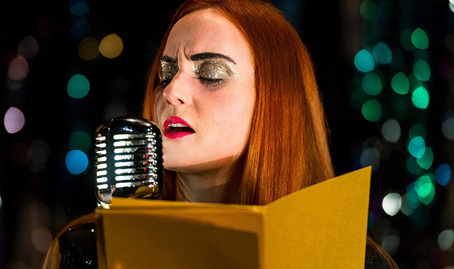 Against a green, glittery background Sandy, wearing gold glittery eyeshadow and bright red lipstick is singing into a classic-style microphone. Whilst she sings she is reading through a pile of letters.