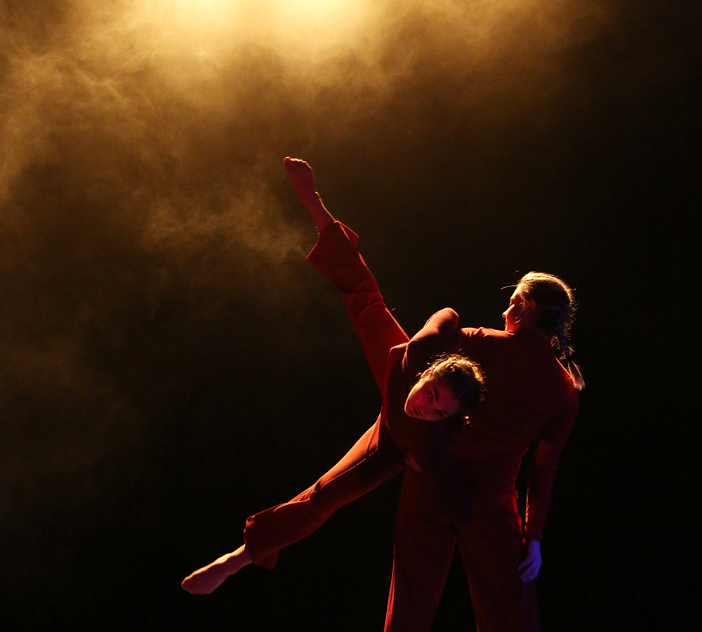 Two performers in a dimly lit, orange spotlight performing 'An Event' choreographed by James Wilton at The New Adelphi Theatre. Both performers are wearing red long sleeved leotards and red trousers. One performer is carrying the other one in the air. 