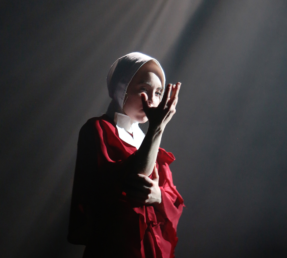 A performer from Emergence is stood in a white spotlight on stage, looking at her hands solemnly. She is wearing a red poncho and white bonnet, similar to the characters from Margaret Atwood's 'The Handmaid's Tale'. 