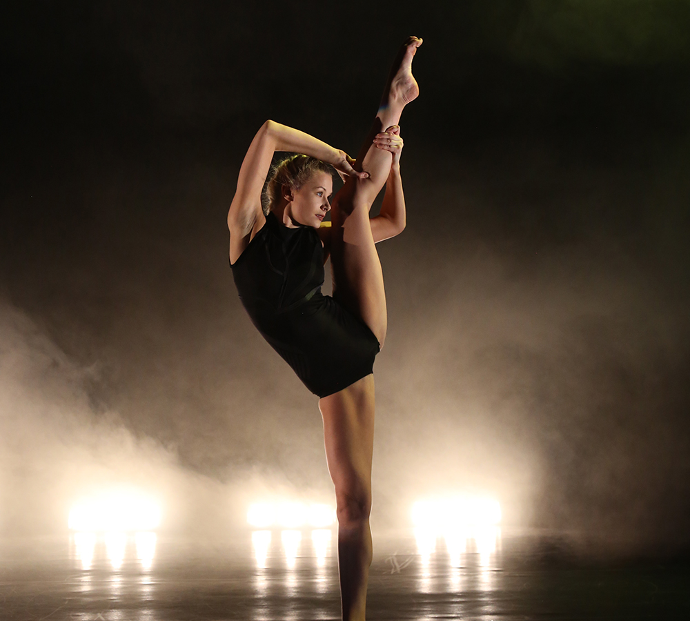 With bright, warm lights in the background of the stage, a performer from 'Emergence' dons a black leotard and is performing a classical ballet move on the stage in which she raises her leg almost above her head.