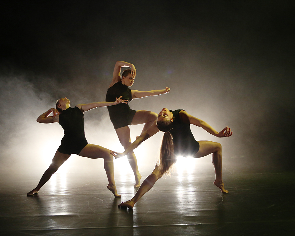 Three dancers from 'Emergence' are on the stage of The New Adelphi Theatre wearing black leotards. Each dancer is in a different pose as they perform in unison on stage.