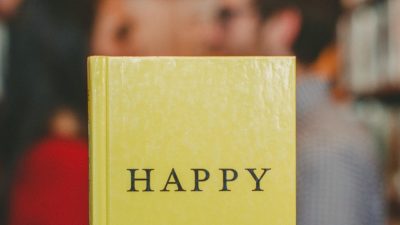 Photograph of a book with "Happy" written on the front cover, held up in front of a couple facing towards each other alongside some bookshelves