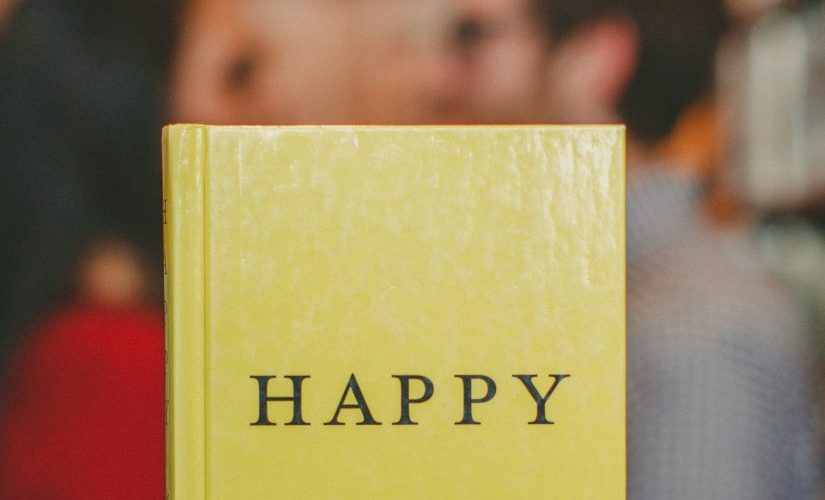 Photograph of a book with "Happy" written on the front cover, held up in front of a couple facing towards each other alongside some bookshelves