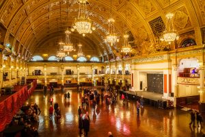 A photo of the Winter Garden's in Blackpool. The Inside of the building is made of white stone and gold paint. There are multiple chandeliers and people are stood waiting.