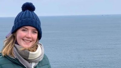 Niamh standing in front of sea. She is wearing a blue bobble hat, green coat and grey scarf.