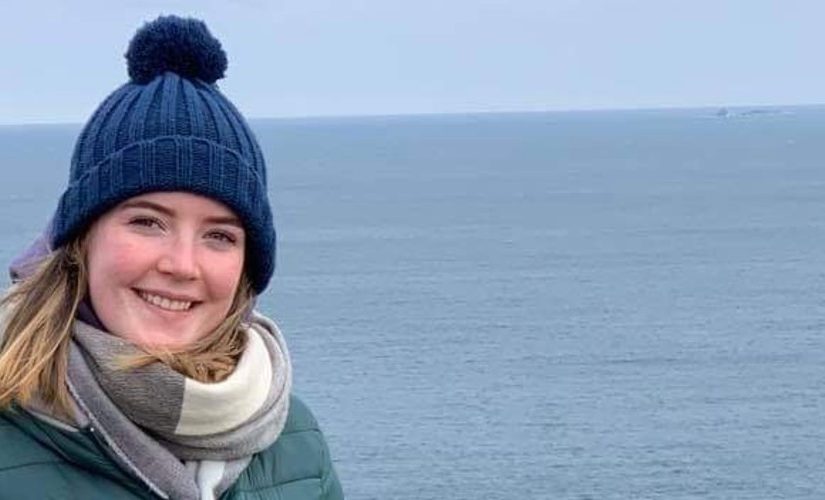Niamh standing in front of sea. She is wearing a blue bobble hat, green coat and grey scarf.