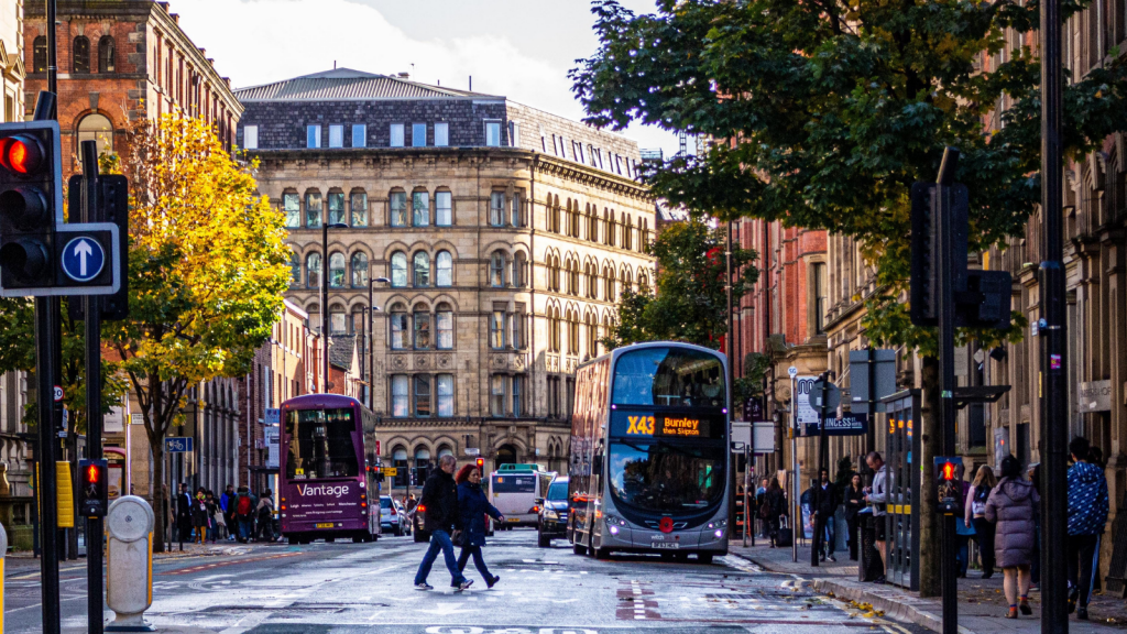 A busy street in Manchester with a couple walking across a road and busses in the background