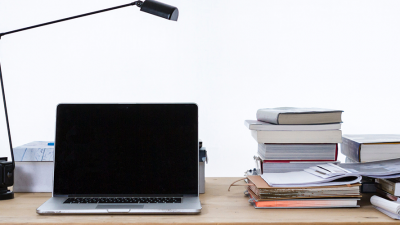 Photo of a latop, with desklamp, books, folder from let to right on top of wooden table