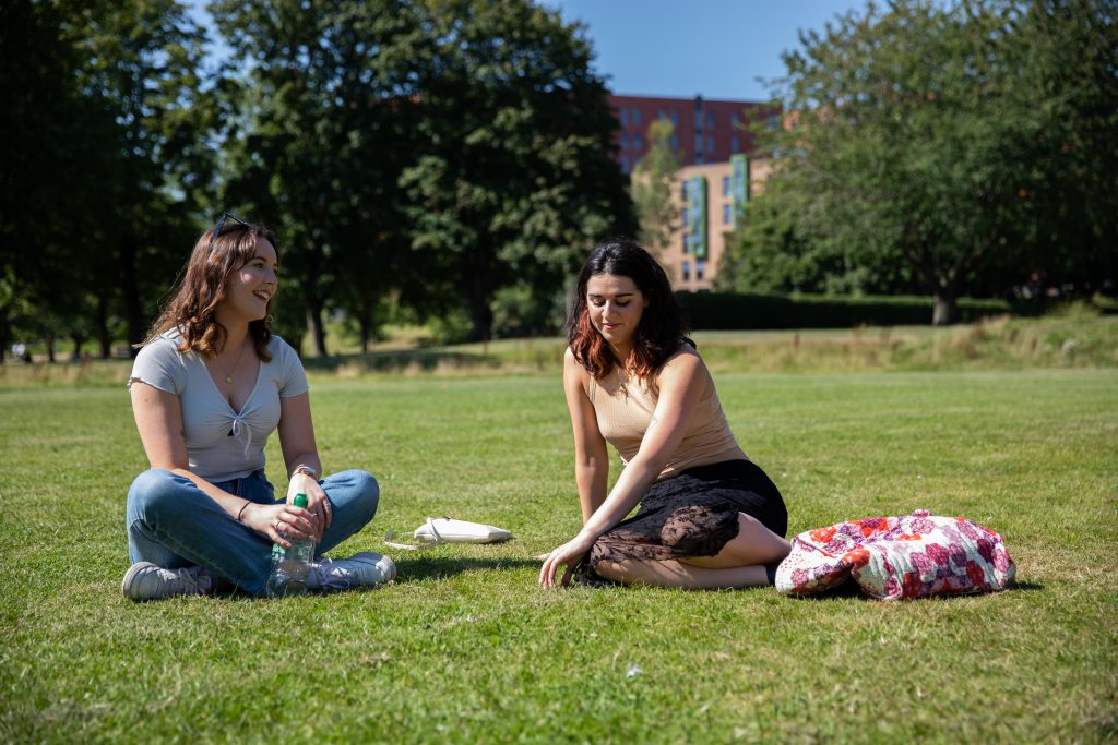 Photograph of two people sat outside on some grass in a park.