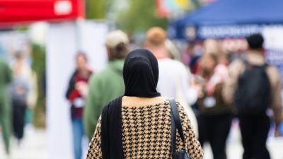 Photograph of Welcome Week at the University of Salford, outside, with a person in a headscarf in the centre of the frame, facing away from the camera. Stalls can be seen in the background.