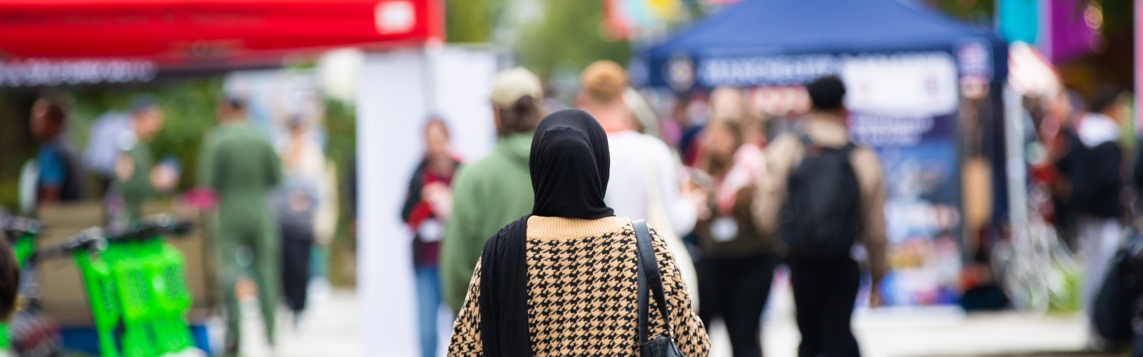 Photograph of Welcome Week at the University of Salford, outside, with a person in a headscarf in the centre of the frame, facing away from the camera. Stalls can be seen in the background.