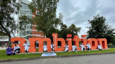 Students in lab coats post next to the 'ambition' sign outside of Salford's Maxwell building