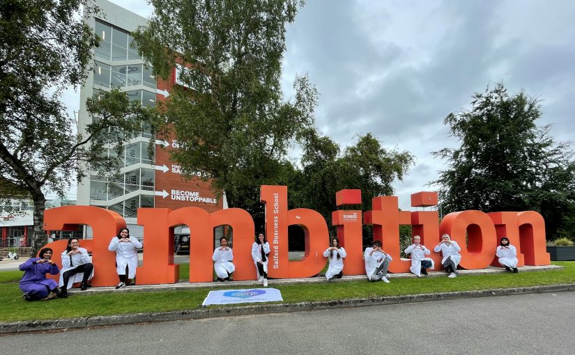 Students in lab coats post next to the 'ambition' sign outside of Salford's Maxwell building