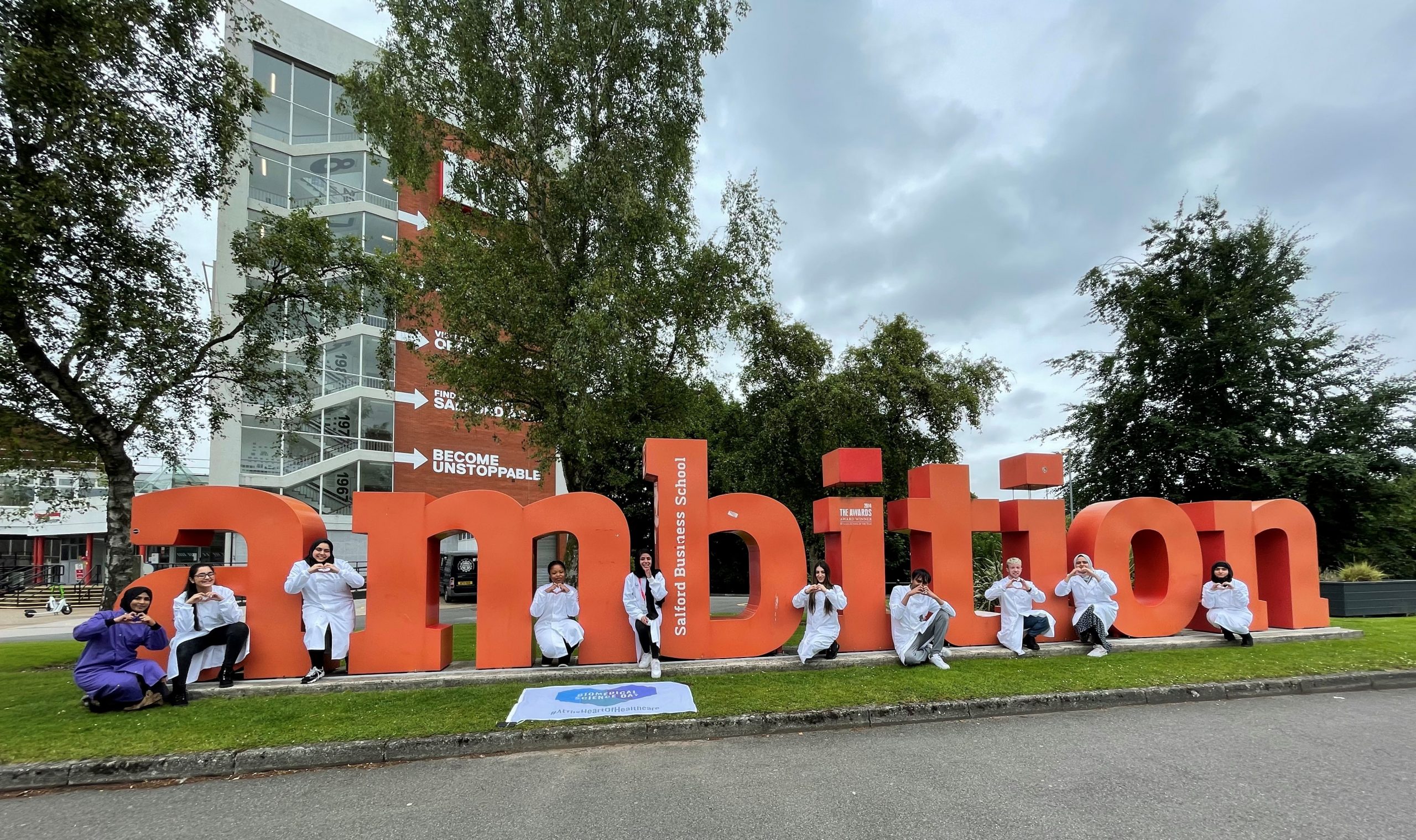 Students in lab coats post next to the 'ambition' sign outside of Salford's Maxwell building