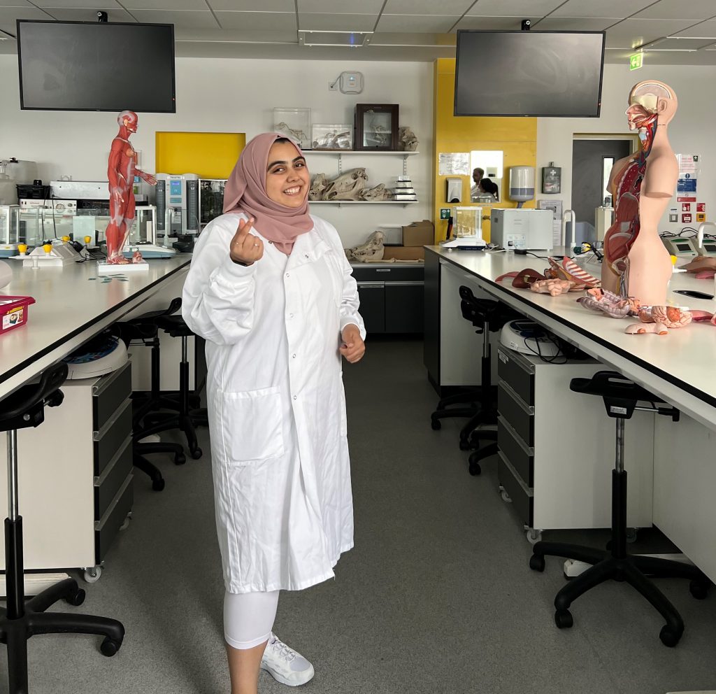 Alia stands in the laboratory, wearing a white lab coat and pink headscarf and is making a heart with her right hand.