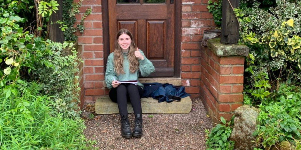 Jas sat on a doorstep, writing in a notebook and giving a thumbs up to the camera