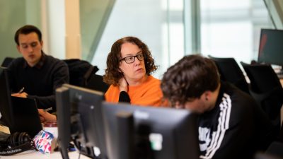 Erica in an orange jumper, sat in front of a computer beside course mates while listening to someone speaking.