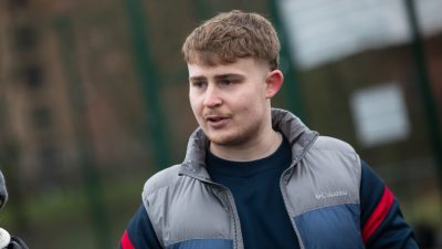 Callum Watts, a young man with light brown hair, is outdoors wearing a blue and grey Columbia jacket. He appears to be in conversation with someone off-camera.