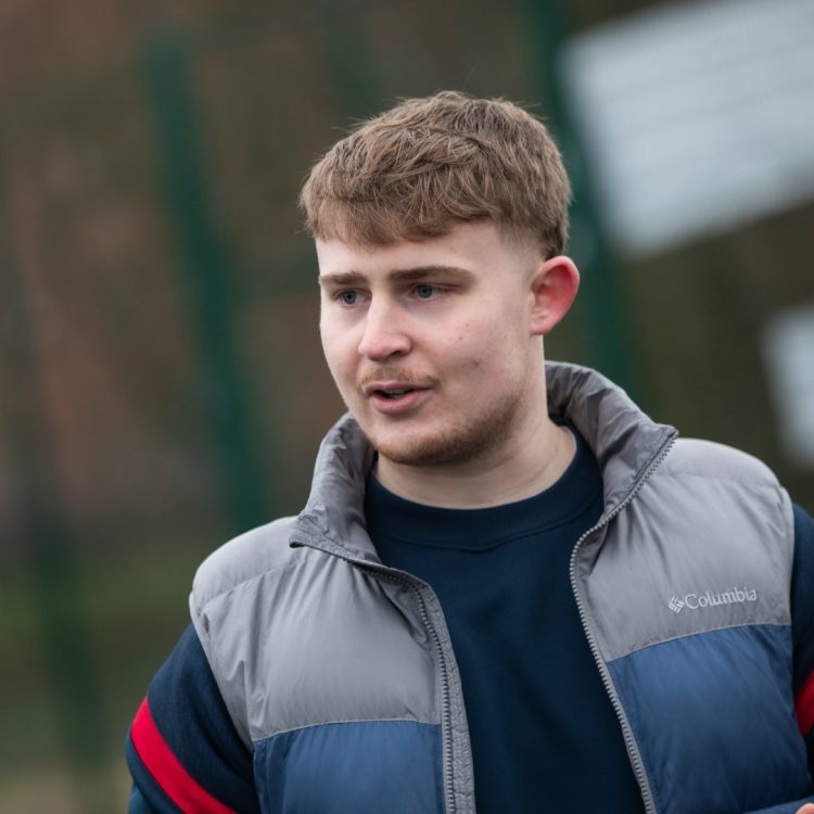 Callum Watts, a young man with light brown hair, is outdoors wearing a blue and grey Columbia jacket. He appears to be in conversation with someone off-camera.