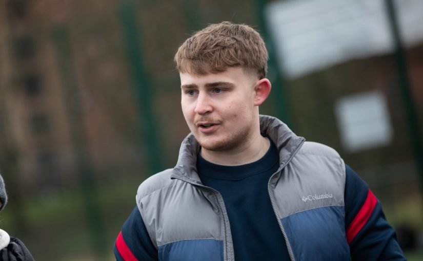 Callum Watts, a young man with light brown hair, is outdoors wearing a blue and grey Columbia jacket. He appears to be in conversation with someone off-camera.