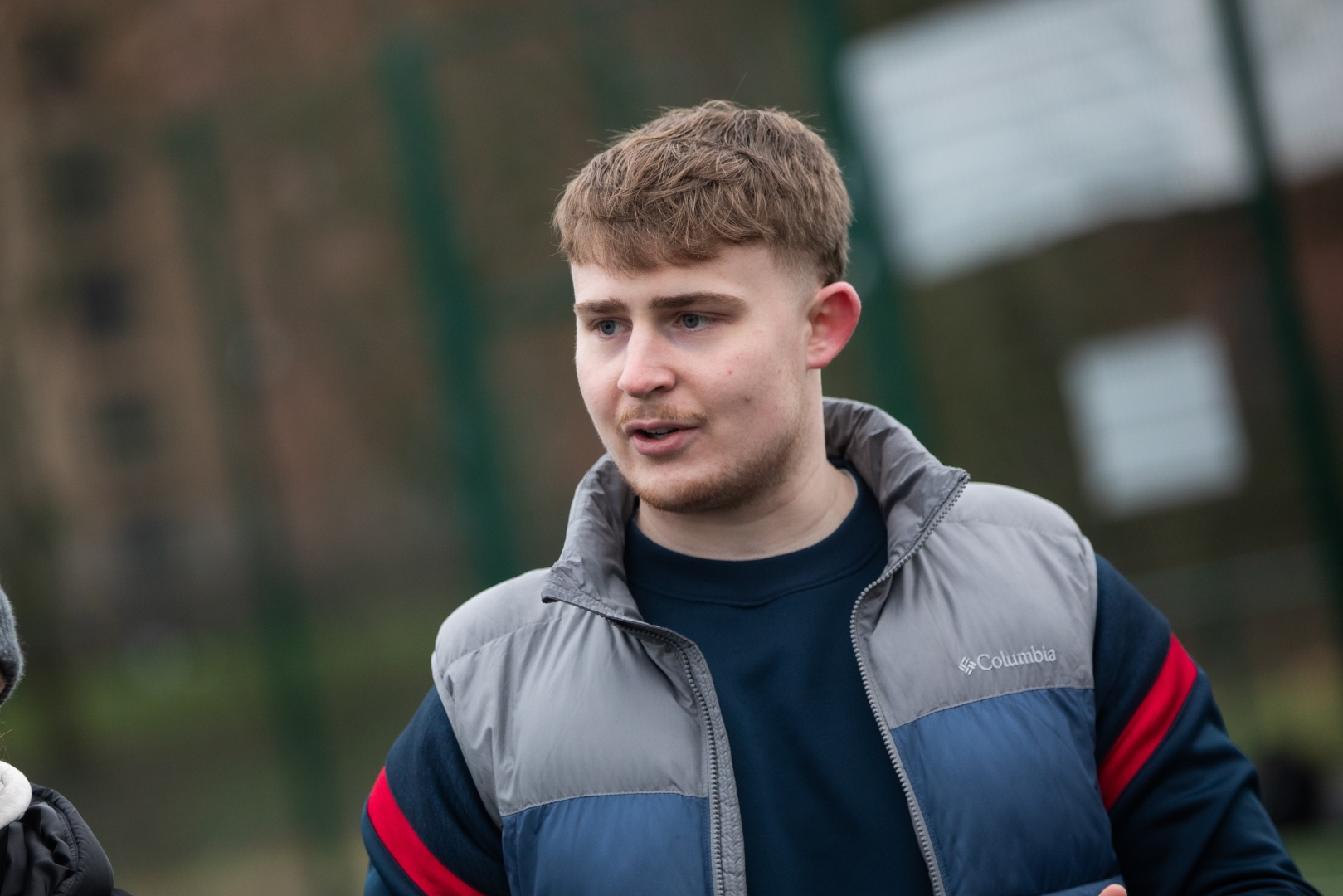 Callum Watts, a young man with light brown hair, is outdoors wearing a blue and grey Columbia jacket. He appears to be in conversation with someone off-camera.