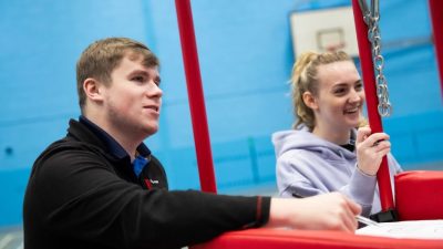 A young man with light brown hair, wearing a black Craghoppers jacket, sitting beside a young woman with blonde hair tied back, wearing a light purple hoodie, both smiling and engaging in conversation next to a red sports structure in an indoor sports facility with blue walls.