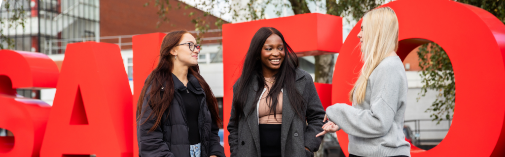 Three students are talking outside the Salford sign.